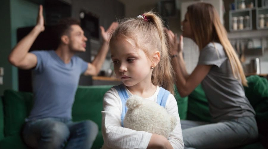 Image of little girl clutching a teddy bear with adults arguing behind her - How Does Childhood Trauma Affect Relationships