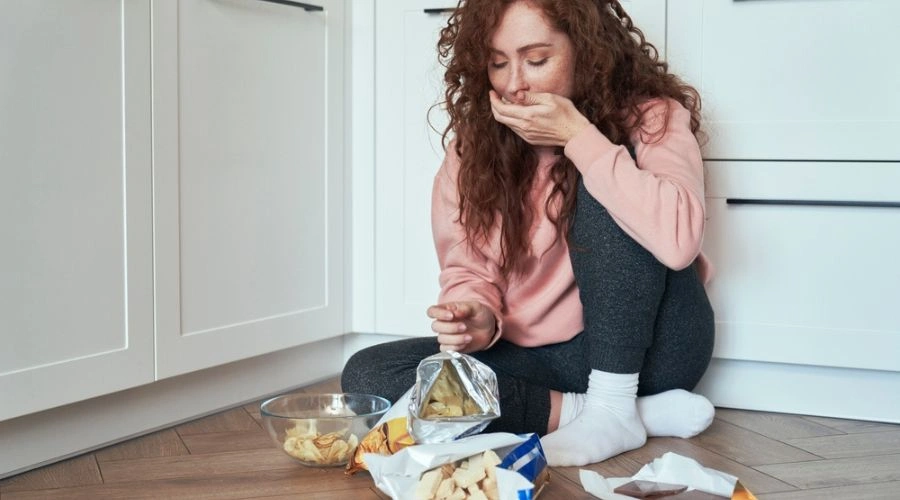 Female sitting with food on the floor, holding hand on mouth - What is a Stress Eating Disorder