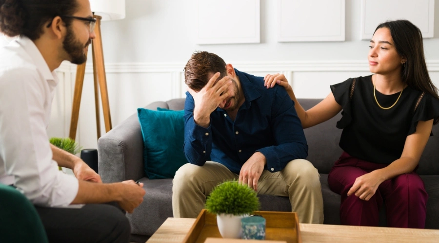 Image of a male sitting on a sofa, beside a female trying to console him as a counselor looks on