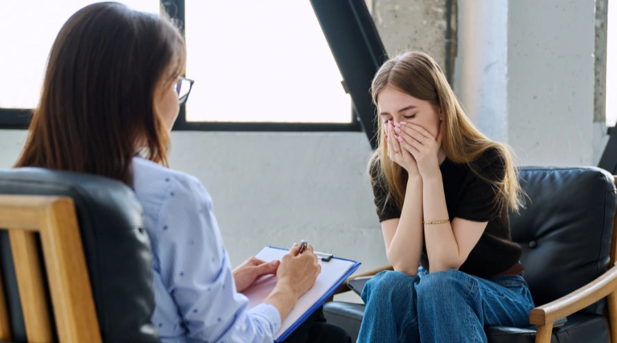 Image of a female sitting in a therapy session with her eyes closed and hands on her face, as her therapist looks on beside her