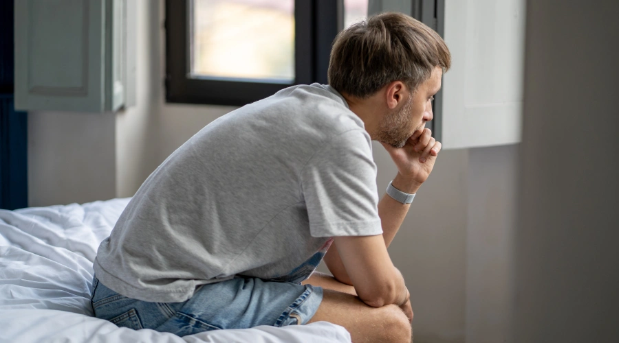 Male sitting along on the front of a bed in deep thought and holding on hand on his chib