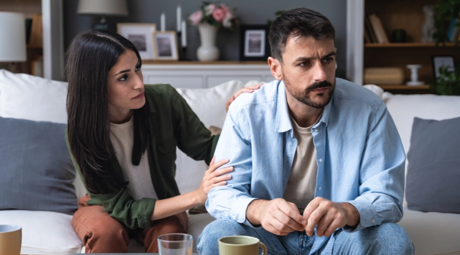 Image of a distraught male sitting on a sofa with a female beside him, holding his shoulder and providing comfort